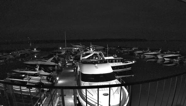 A nighttime view of a marina filled with various boats and yachts docked along a wooden pier. The scene is predominantly dark, with limited illumination highlighting the shapes of the boats. Several boats are tied up closely together, and the water reflects faint lights from the vessels. The perspective suggests a vantage point from a higher position, looking out over the marina.