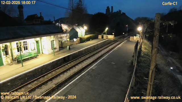 A dimly lit railway station at dusk. The platform features a long, low stone wall on one side and a green wooden bench with simple metal legs. An old building with a stone façade and green details is visible, displaying several windows illuminated by warm light. There is a sign on the platform indicating "WAY OUT." The railway tracks extend into the distance, with a wooden fence bordering the platform. In the background, silhouettes of hills and a castle-like structure are faintly visible against the darkening sky. The atmosphere appears quiet and serene, typical of a rural station at twilight.