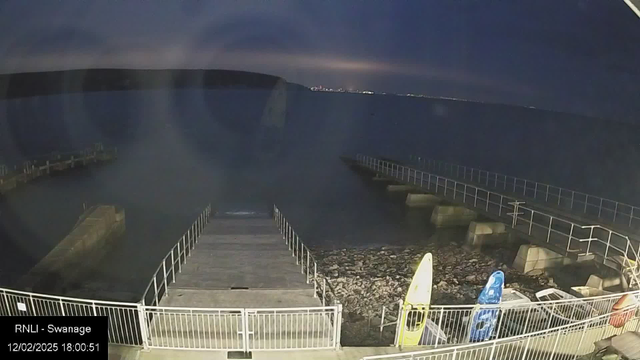 A dimly lit scene of a waterfront area at dusk. In the foreground, there is a wooden ramp leading down to the water, flanked by metal railings. To the right, there are two colorful kayaks, one yellow and one blue, resting on a rocky shore. The water is calm and extends towards the horizon, where faint lights from a distant shoreline are visible. The sky is partially cloudy, with shades of blue and gray indicating twilight. A small section of a pier is visible on the far left, and the scene captures a tranquil evening atmosphere.