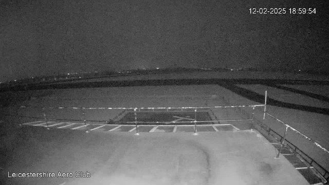 A dimly lit image from a webcam showcasing an airstrip. In the foreground, there is a railing along a flat surface with white lines indicating parking spaces. The background features a dark sky with a faint outline of distant lights or structures. The overall scene appears quiet and undisturbed, conveying a nighttime atmosphere.