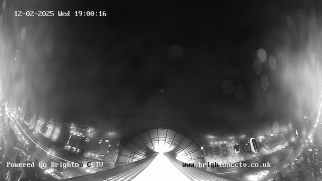 A black and white webcam image showing a view from a high vantage point overlooking a city at night. The foreground features a brightly lit pathway or road leading into a dark tunnel. Buildings and structures are visible on both sides, with lights reflecting off wet surfaces, creating a glistening effect. The timestamp at the top indicates it is February 12, 2025, at 7:00 PM, and there are some circular light blurs from lens flare in the upper part of the image. The bottom left shows "Powered By Brighton CCTV" and the website URL.