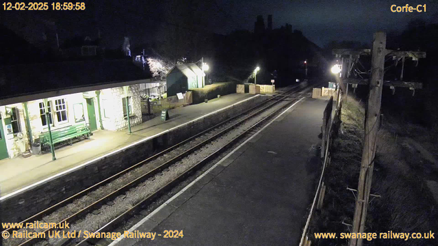 A dimly lit train station platform at night. On the left, there is a stone building with large windows and a green bench outside. A green sign near the platform reads "WAY OUT." The platform is bordered by wooden fencing, and a nearby street lamp casts a faint light. The railway tracks run parallel, leading into darkness, while a wooden pole with wires stands on the right. In the background, shadowy outlines of trees and hills are visible.