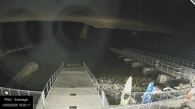 A nighttime image of a harbor scene. In the foreground, there is a concrete walkway leading out over the water, bordered by a railing. To the right, a small area of rocky shore is visible, along with several colorful surfboards resting against the railing. The water is dark and undulating, reflecting faint lights from a distant coastline in the background. The sky is mostly dark, with some cloud cover obscuring the stars. The image is somewhat grainy, typical of low-light conditions.