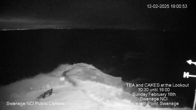 A dark, nighttime scene featuring the ocean with waves crashing against a rocky shoreline. A small sign is visible on the left side of the image. The horizon is barely illuminated by distant light, suggesting the presence of land or buildings in the far background. A bench is situated near the edge of the rocks, and the overall atmosphere feels tranquil and remote. There is text in the lower left corner indicating the source of the image as "Swanage NCI Public Camera," along with event information on the right side.