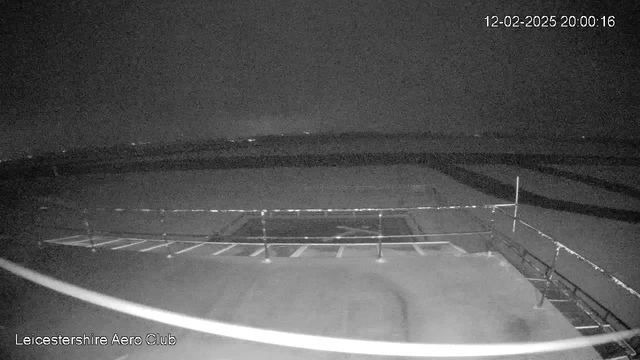 A dark scene captured from a webcam at the Leicestershire Aero Club. The image is primarily black with minimal visible details. In the foreground, there is a partially visible tarmac area with several white parking lines. A slight outline of a structure, possibly a railing or fence, can be seen towards the right side of the image. In the background, faint lights suggest distant buildings or vehicles, but the overall visibility is low due to the darkness. The timestamp shows it is 8:00 PM on February 12, 2025.