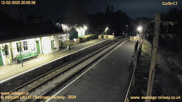 A dimly lit railway station platform at night. To the left, there is a stone building with large windows, a green bench, and a sign partially visible. The platform is empty, with two railway tracks running along it. On the right, a wooden fence marks the exit, and there are several lights illuminating the area. In the background, a hilly silhouette can be seen against the night sky, and a few trees are also visible. The atmosphere is quiet and deserted.