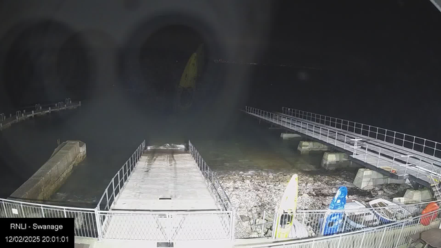 An image of a dark waterfront scene at night, featuring a partially visible pier extending into calm water. To the right, a few brightly colored kayaks are stored on the shore, with a yellow kayak prominently displayed. The surrounding area shows rocky terrain and a low fence alongside the pier. The water near the shoreline is slightly rippling under the moonlight, with faint lights visible in the distance across the water. The date and time in the corner indicate it is February 12, 2025, at 20:01:01.