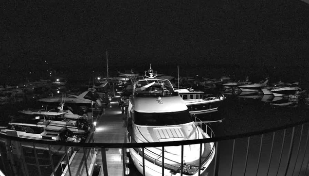 A dimly lit marina at night, featuring multiple boats docked closely together. In the foreground, a large white yacht is prominently positioned, with its sleek hull reflecting faint light. The wooden walkway leading from the viewer’s perspective narrows between the boats, creating a sense of depth. The surrounding boats are mostly dark with some illuminated spots, suggesting a serene nighttime atmosphere on the water.