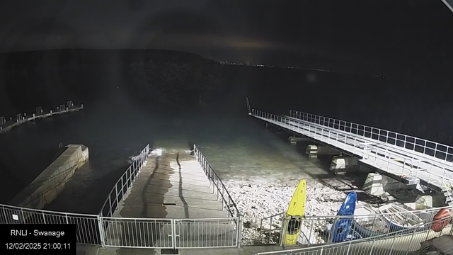 A dimly lit scene at a waterfront at night. In the foreground, a concrete ramp leads down to the water, bordered by a white railing. To the right, there are two kayaks: a yellow one and a blue one, resting on the shore. In the background, a long wooden pier extends into the water, with several posts visible at the end. The water appears calm, and there's a dark shoreline in the distance, barely illuminated by ambient light. The date and time display on the bottom left corner reads "RNLI - Swanage 12/02/2025 21:00:11."