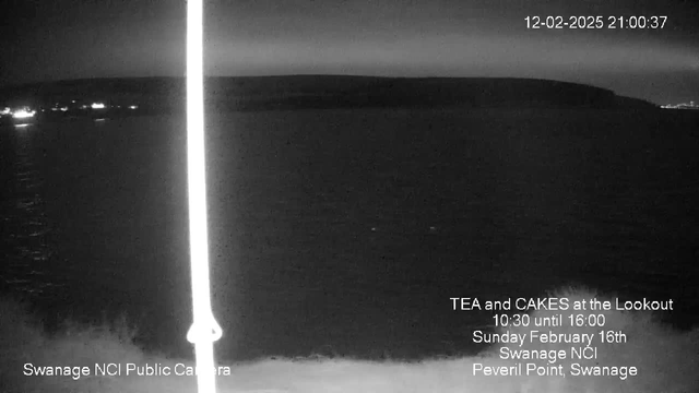 A dimly lit seaside view at night, with a calm, dark sea merging with the horizon. A faint light glimmers off the water, suggesting distant lights from a shore. The image features a vertical white line, likely a reflection or glare. In the lower part of the image, text indicates this is a webcam from Swanage NCI, advertising an event for tea and cakes on February 16th, 2025.