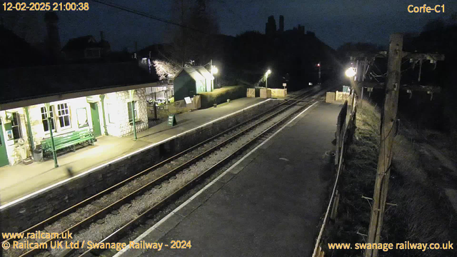 A dimly lit railway station scene at night. The station building, made of stone, has green accents and features illuminated windows. A green bench is visible on the platform. There are two illuminated lampposts along the platform, casting light on the gravel and concrete. The railway tracks run through the scene, leading into the distance. A wooden fence borders the platform, with a sign indicating "WAY OUT" visible. In the background, silhouettes of trees and possibly a hill are faintly illuminated. The atmosphere is quiet and tranquil, typical of a rural station at night.