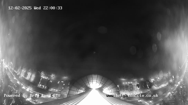 A black and white webcam view showing a cityscape at night. The top portion displays a bright light source, possibly a building or structure, illuminating the scene. Below, roadways with faint light reflections can be seen, suggesting vehicle movement. The image has a blurred and hazy quality, indicating possible moisture on the camera lens. A timestamp indicating the date and time is in the upper left corner, along with a logo in the lower left.