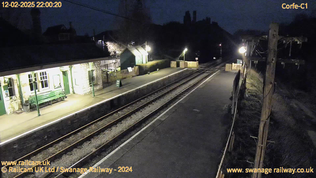 A dimly lit train station at night. The platform features a green bench and several signs. To the left, there is a stone building with windows, likely the station house. The right side shows two railway tracks leading into darkness, with wooden picket fences along the edge of the platform. An electric pole with wires stands on the far right. A green sign on the platform reads "WAY OUT."