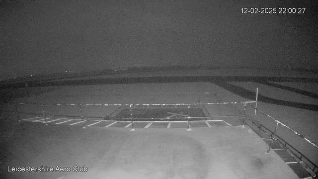 A dimly lit scene of an airfield at night. The asphalt area is mostly dark with a few white lines marking parking spaces. A low railing is visible in the foreground. In the distance, faint lights indicate the presence of buildings or vehicles. The image is captured from a webcam at the Leicestershire Aero Club, with the date and time displayed in the top right corner.