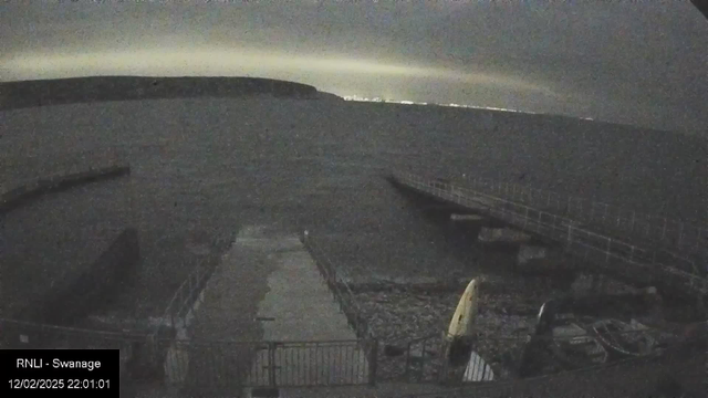 A dark waterfront scene featuring a rocky shoreline and a wooden pier extending into the water. The sky is cloudy with limited light, creating a moody atmosphere. A few small vessels are visible at the edge of the pier. In the background, a faint outline of hills can be seen on the horizon, along with blurred lights suggesting a distant shoreline. The time and location are noted at the bottom left of the image.