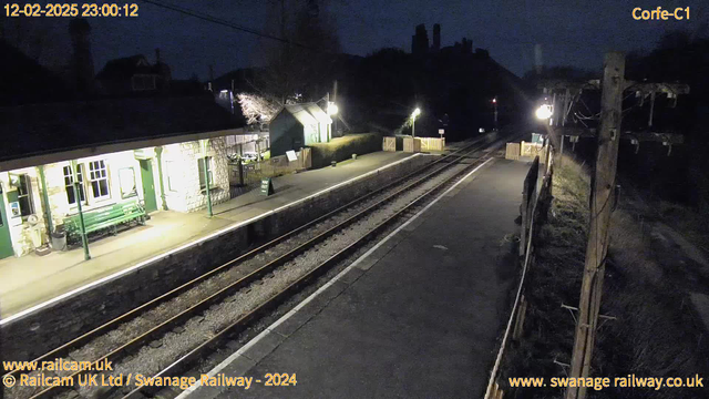 A dimly lit railway station platform at night, featuring a stone building with green detailing and a green bench. On the left, there are small illuminated signs and a wooden fence. The platform is lined with tracks leading off into the darkness. In the background, there are trees and silhouettes of buildings, partially illuminated. A single light illuminates part of the scene, creating a serene but quiet atmosphere.