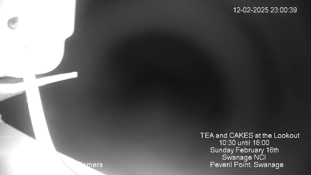 A black and white webcam image showing a blurred background, with slight light reflections on the left side. Text in the lower right corner provides event details: "TEA and CAKES at the Lookout, 10:30 until 16:00, Sunday February 16th, Swanage NCI, Peveril Point, Swanage." The upper right corner shows the date and time: "12-02-2025 23:00:39."