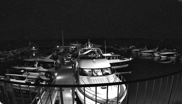 A dimly lit marina at night, featuring several boats docked along a wooden pier. The scene is illuminated by sparse light from surrounding areas, casting reflections on the water. A few boats are larger yachts, while others are smaller vessels. A railing is visible in the foreground, framing the view of the marina and the boats. The overall atmosphere is tranquil but dark, typical of a nighttime setting.