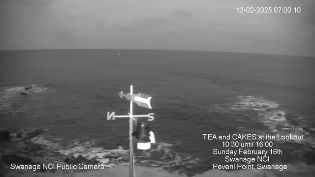 A webcam view of a calm sea at dawn, showing a light gray horizon where the water meets the sky. In the foreground, there's a black and white weather vane with directional indicators (N, S, E, W) prominently displayed. The bottom of the image includes text advertising a tea and cake event, with the date and location details noted. The scene conveys a tranquil atmosphere, with gentle waves lapping at the shore.