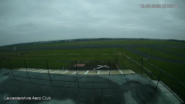 A view from a rooftop overlooking a wide, grassy airfield. The sky is overcast and gray. In the foreground, there are railings and part of a flat rooftop. Below, a landing area is marked with a large white "X" on the ground, surrounded by a fence. In the distance, the airfield extends to the horizon, with a few paths visible alongside grassy areas.