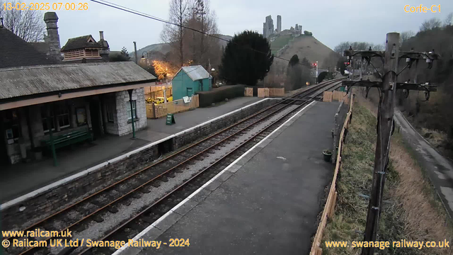 The image shows a railway station with a stone platform and wooden fences along the right side. There are two railway tracks visible, leading into the distance. On the left, a building with a slanted roof is visible, featuring large windows and a green bench. A blue-green shed with a sign is situated nearby. In the background, there are hills with ruins of a castle atop one, partially obscured by trees. The scene is set during early morning with a cloudy sky.