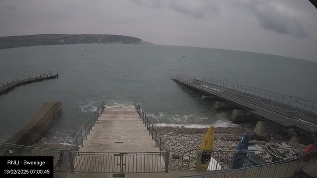 A cloudy scene at a seaside location featuring a concrete ramp leading down to the water. To the left, there is a pier extending into the sea, with small waves visible. The shoreline consists of pebbles, with several colorful boats stored near the ramp, including a yellow kayak. The backdrop shows cliffs in the distance under the overcast sky.