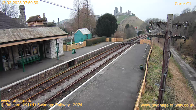 A view of Corfe Castle railway station with a stone platform and wooden fence. To the left, there is a green bench under a roof with sloping tiles. In the background, there is a blue shed and a sign indicating "WAY OUT." The railway tracks run parallel to the platform leading into the distance. On the right, a wooden utility pole stands with electrical wires. In the background, Corfe Castle is visible on a hill, surrounded by trees. The sky is overcast.