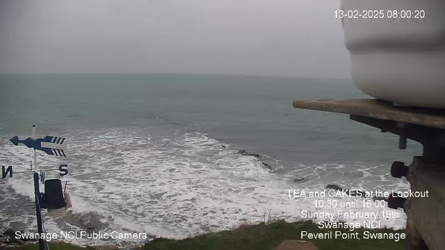 A cloudy, overcast sky is visible above a choppy sea with white-capped waves. In the foreground, there is a weather vane displaying the cardinal directions, with the letters 'S' for south. A rocky shoreline is partially seen at the bottom of the image, with grass and small plants growing nearby. The camera view captures the tranquil yet tumultuous beauty of the coastal landscape. Text overlays provide event details and the camera location.