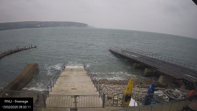 A view of a coastal area featuring a wide expanse of gray water under a cloudy sky. In the foreground, there is a set of concrete steps leading down to the water, bordered by a metal railing. To the sides, there are wooden piers extending out over the water, with several boats lined up, including a yellow kayak and blue and red boats. Along the edges, there are rocky formations and scattered stones. The distant shore is visible, with green hills and cliffs fading into the misty background.