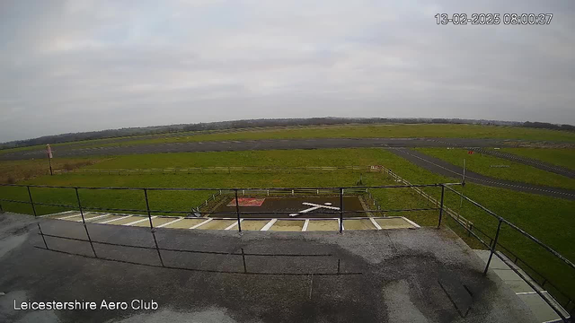 A view from a webcam showing a grassy airfield under a cloudy sky. In the foreground, there is a railing with a flat rooftop surface below it. The airfield features a large runway and a helipad marked with a white cross. There are fields and a fence surrounding the area, and some signs in the distance. The image is taken during early morning hours, as indicated by the timestamp in the corner.