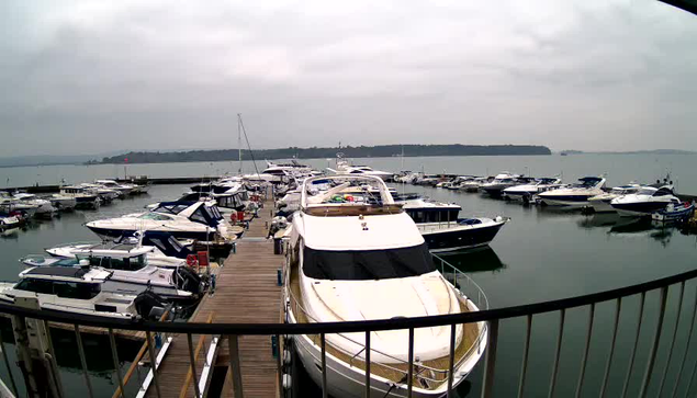 A marina scene featuring numerous boats docked along a wooden pier. The foreground shows a prominent white yacht with a large cabin and multiple small boats around it. The water is calm and reflects the cloudy sky above. In the background, a green hillside is partially visible on the horizon, hinting at an overcast day.