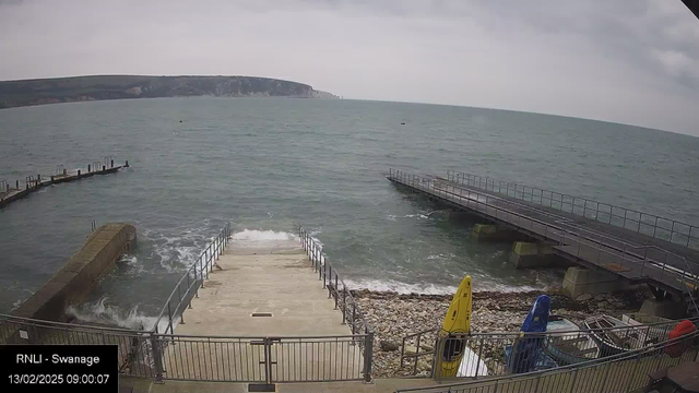 A view of a coastal area with a cloudy sky. In the foreground, a concrete staircase leads down to the water, flanked by a metal railing. To the left is a stone wall and a curved wooden pier extending into the sea, with several posts visible. On the right, a smaller wooden platform juts out, and various colorful kayaks, including yellow and blue, are placed near the water's edge. The sea appears choppy with small waves, and a distant cliff can be seen in the background.