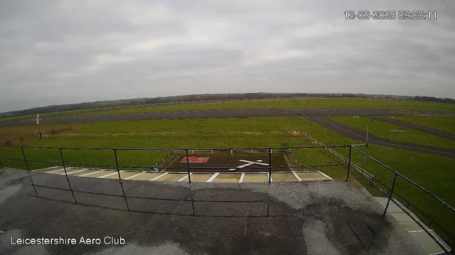 The image shows a view from a raised platform at the Leicestershire Aero Club. In the foreground, there is a railing and a grey surface. Below, a grassy area is visible, along with a small section of pavement. A white X marking is on the ground, indicating a possible stopping point for aircraft. Beyond the grassy area, there is a black runway extending to the horizon. The sky is overcast with grey clouds, and the landscape in the background includes more fields and a distant tree line.