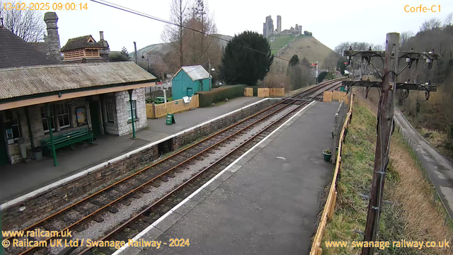 A view of Corfe Castle railway station. In the foreground, there is a stone platform with tracks leading off to the right. A green wooden bench is situated on the platform, alongside a poster on the wall. To the left, there is a brick building with a red-tiled roof, and beyond it, a small green shed. A wooden fence encloses the area to the left, and a path leads away from the station. In the background, Corfe Castle is visible on a hill, partially obscured by trees, with a cloudy sky above. The scene appears tranquil and empty.