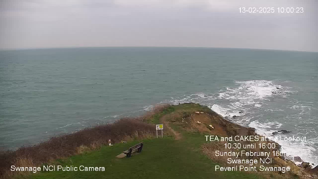 A coastal view showing a grassy cliff with a bench overlooking the sea. The water is a deep blue-green color, with white waves breaking along the rocky shore. The sky is overcast with gray clouds. In the foreground, there are signs indicating an event for tea and cakes, and the area appears serene with sparse vegetation on the cliff.