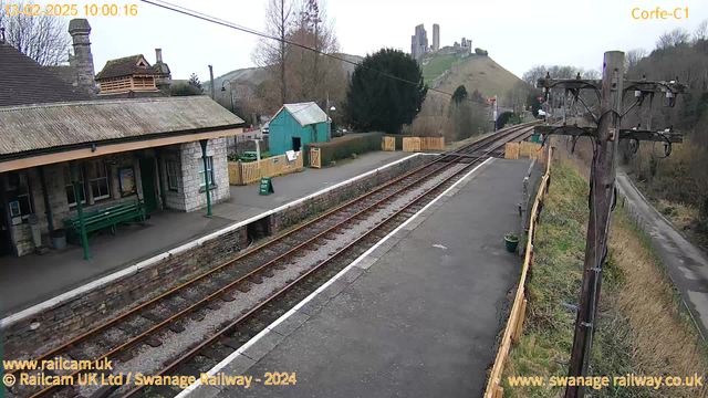 A train station scene featuring a stone platform with a green bench and a wooden structure with a sloped roof, likely a ticket office. Behind the station, there is a small blue building and a wooden fence. In the background, a hill topped with castle ruins is visible, surrounded by trees. A power pole with wires stands to the right. The ground is mostly clear, with railway tracks running along the platform. The sky is overcast, indicating a cloudy day.