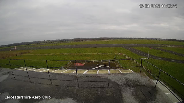 A wide view of an airfield with a grassy area and a runway in the background. The sky is overcast. There are white markings on the ground, including an X shape, and a railing at the foreground. A few trees and a strip of road can be seen in the distance, along with some signs near the perimeter of the field.