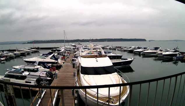 A marina filled with various boats and yachts is visible. There are numerous white and blue vessels docked along a wooden pier. The sky is overcast with gray clouds, and the water reflects the boats and the cloudy sky. In the background, a wooded shoreline can be seen in the distance. The image appears to be taken from a raised vantage point, showing a wide view of the harbor.