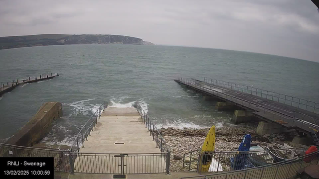 A view of a coastal area featuring a set of concrete steps leading down to the water. On the left, a rocky edge juts into the sea, and in the background, there are green hills and cliffs. In the foreground, two kayaks, one yellow and one blue, are positioned on the shore near some boats. A jetty extends into the water on the right, with some railings visible. The sea appears slightly choppy, and the sky is overcast. The timestamp shows the image was taken on February 13, 2025, at 10:00:59.