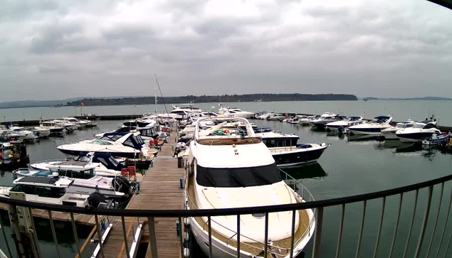 A marina filled with numerous boats docked in calm water. In the foreground, a large white yacht with a blue stripe is prominent, surrounded by smaller boats of various sizes and colors. The scene is set under a cloudy sky, with a distant shoreline visible in the background, framed by a wooden dock and metal railing in the lower part of the image.