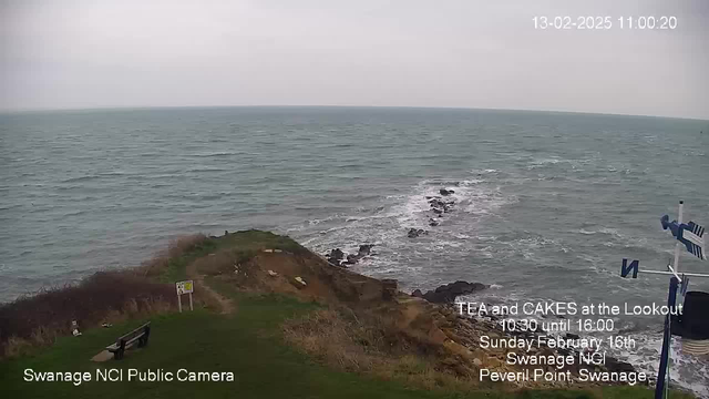 A coastal scene featuring rolling waves in the ocean, with rocky outcroppings visible in the water. The sky is overcast and gray, indicating a cloudy day. In the foreground, a grassy area slopes down towards the water, with a wooden bench positioned to the left. There is a sign nearby, and a directional arrow appearing on a pole to the right, which may indicate nearby attractions. The atmosphere feels calm and serene, typical of a coastal environment.