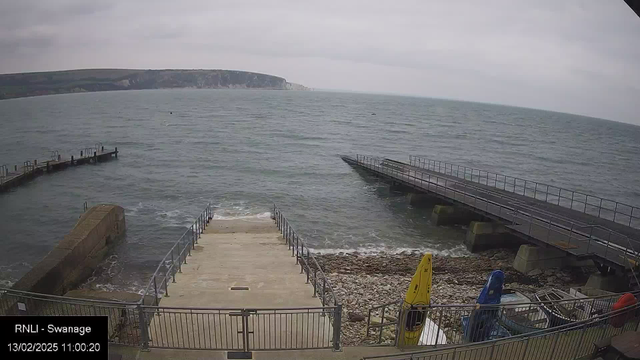 A coastal scene showing the shoreline and sea. In the foreground, there are stone steps leading down to the water, and a railing surrounds the area. To the left, a rocky pier extends into the sea, and on the right, a wider platform or pier is visible. Various colored kayaks—red, blue, and yellow—are positioned near the water's edge. The sky is overcast, with gray clouds covering the horizon, and waves are gently lapping at the shore. The landscape in the background includes a cliff and additional landforms.