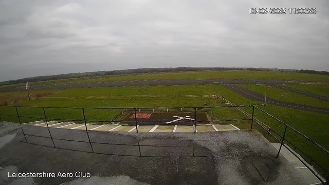 A cloudy scene captured from a webcam at the Leicestershire Aero Club. The foreground shows a railing and a gray surface. In the background, there is a large open field with patches of green grass, intersected by a dark asphalt runway. A small white X marking is visible on the ground, and there are scattered fences along the perimeter of the field. The sky appears overcast, with soft, diffuse light illuminating the scene.