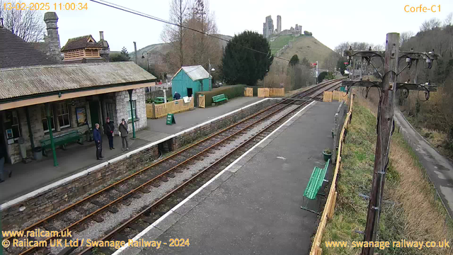 A view of Corfe Castle railway station on a cloudy day. The station features a stone building with a sloped roof, and there are several green benches along the platform. A few people are standing near the entrance of the station, and a green wooden shed is visible in the background. To the left, a train track runs parallel to the platform, leading into the distance. The hill in the background is dominated by the ruins of Corfe Castle, which is partially obscured by trees. A wooden post with electrical wires stands on the right side of the image.