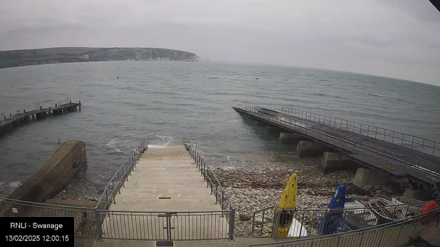 A view of a coastal area featuring a pier extending into the water on the left side. In the foreground, there is a wide concrete ramp leading down to the water. To the right, several colorful kayaks are placed on the ground, with a yellow kayak prominently visible. The sea is calm with gentle waves, and a rocky shore is seen along the bottom. The sky is overcast with gray clouds.