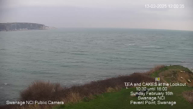 A coastal view featuring a gray, overcast sky. The sea is visible, with gentle waves disturbing the surface. In the foreground, there is a patch of grassy land with some low bushes, and a portion of the cliffside can be seen to the left. On the right, signs indicate a lookout area. The time and date are displayed in the top corner of the image.