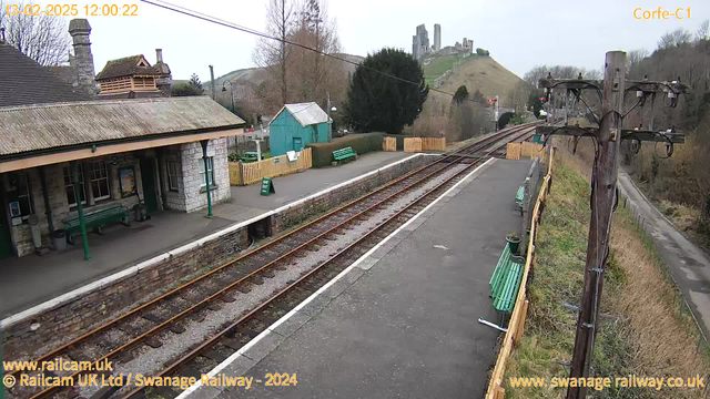 A view of Corfe Castle railway station featuring two railway tracks extending into the distance. To the left, there is a stone building with a slate roof, and a green bench along the platform area. A wooden fence encloses the station with a small, turquoise building visible in the background. On the hillside behind the station, ruins of Corfe Castle are visible, surrounded by trees. The sky is overcast, indicating a cloudy day.