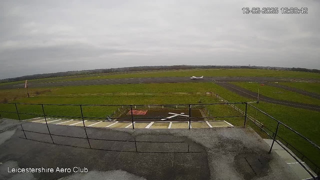 A webcam view of an aerodrome shows a large grassy field with a runway and a light aircraft on the right side of the image, taxiing on the tarmac. The sky is overcast with clouds, and there is a railing visible in the foreground. In the background, there are distant hills and trees. The scene appears to be calm and quiet, with no visible activity other than the aircraft. The lower part of the image has a partial view of a flat rooftop.