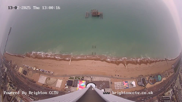 Aerial view of a beach with a sandy shore and gentle waves lapping at it. The ocean is a greenish-blue, and there is a large pier structure partially submerged in the water. The beach is lined with various structures, including a boardwalk and colorful entertainment venues. The image is taken from a high vantage point, looking straight down, with the date and time displayed in the corner.