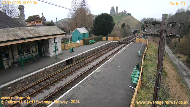 A view of a railway station with a stone building featuring green benches under an awning. There are railroad tracks running alongside the platform. In the background, a hill is visible with castle ruins atop it, surrounded by trees and a cloudy sky. A green shed and a sign indicating "Way Out" are also present. An electrical pole with several wires stands on the right side of the image.